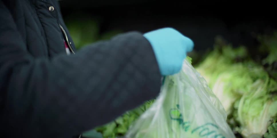 A woman wearing gloves and a mask places lettuce in a plastic bag in the produce section of the grocery store