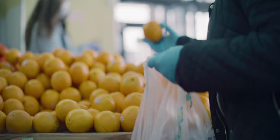 A shopper wearing gloves selects oranges from a grocery store display and places them in a plastic bag