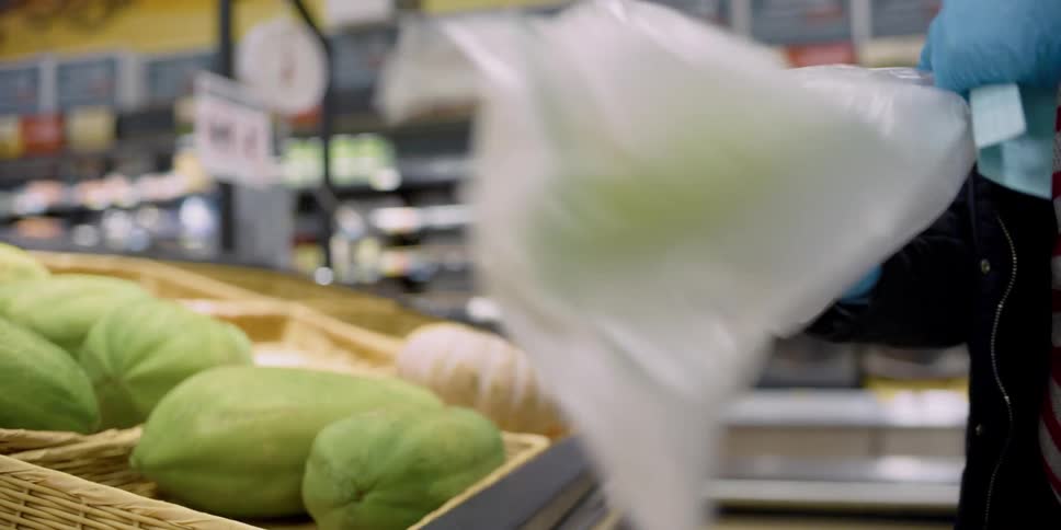 A woman chooses a melon at a grocery store and places it in a plastic bag