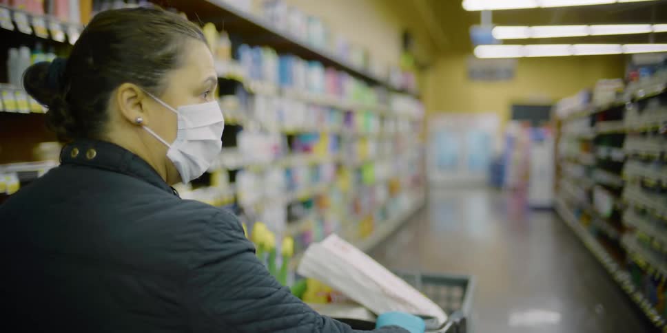 A woman wearing a mask and gloves pushes a shopping cart down the aisle of a store
