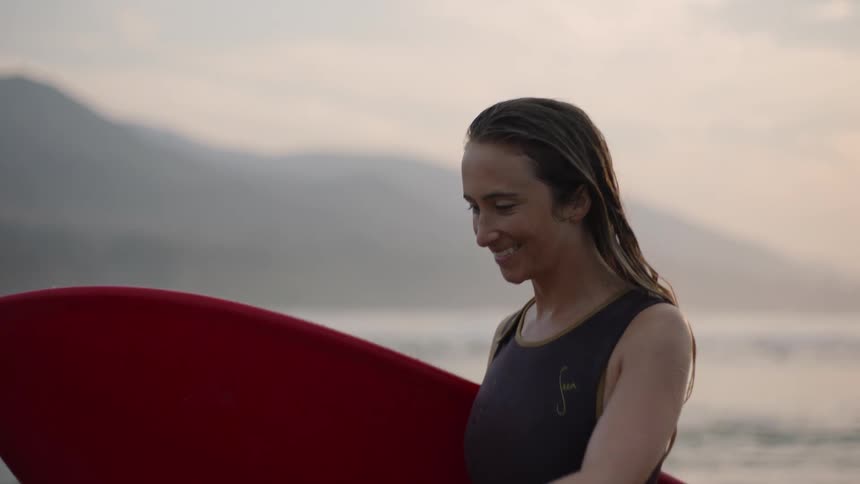 A smiling woman carries a surfboard along a beach
