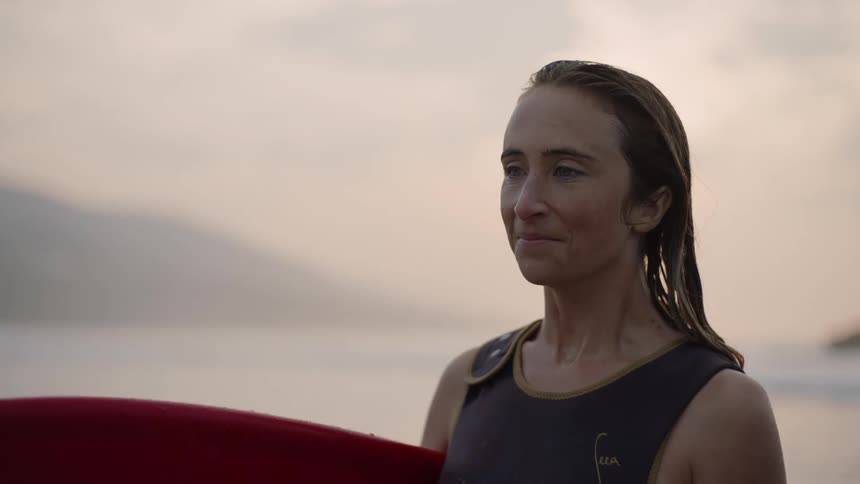 A woman is standing on the shore holding her surfboard as she smiles and turns her head