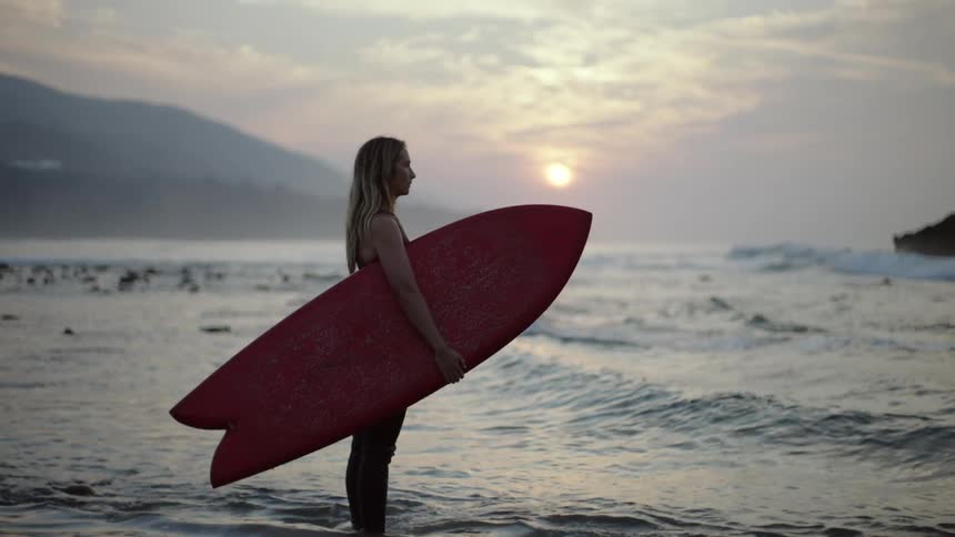 A woman stands in the low tide on the beach while holding a surf board in the evening