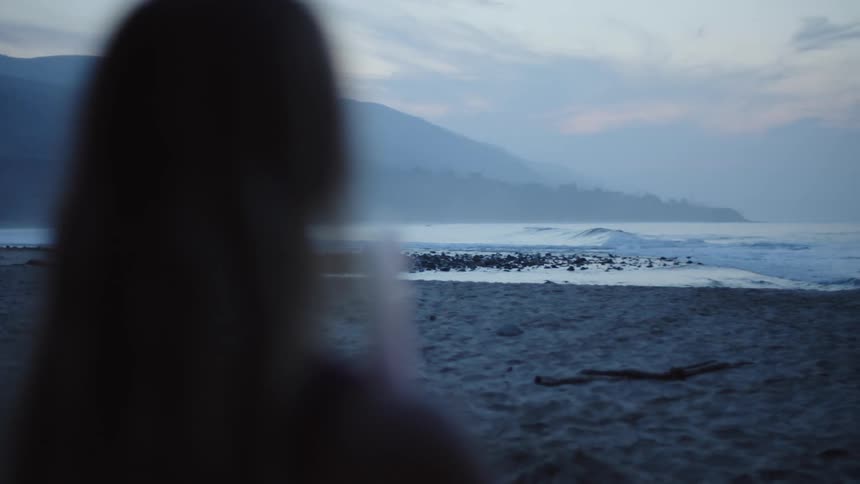 Woman with brown hair holds surfboard stands on the beach holding surfboard at dusk