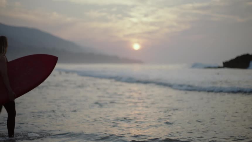 Female surfer in black wetsuit carries surfboard into sea at sunset