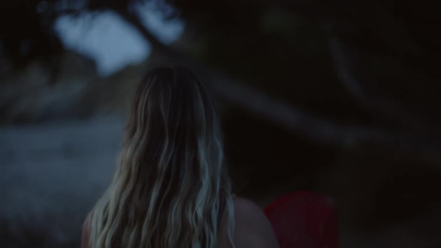 The backside of a girl with blonde hair walking with something red under her arm toward a beach