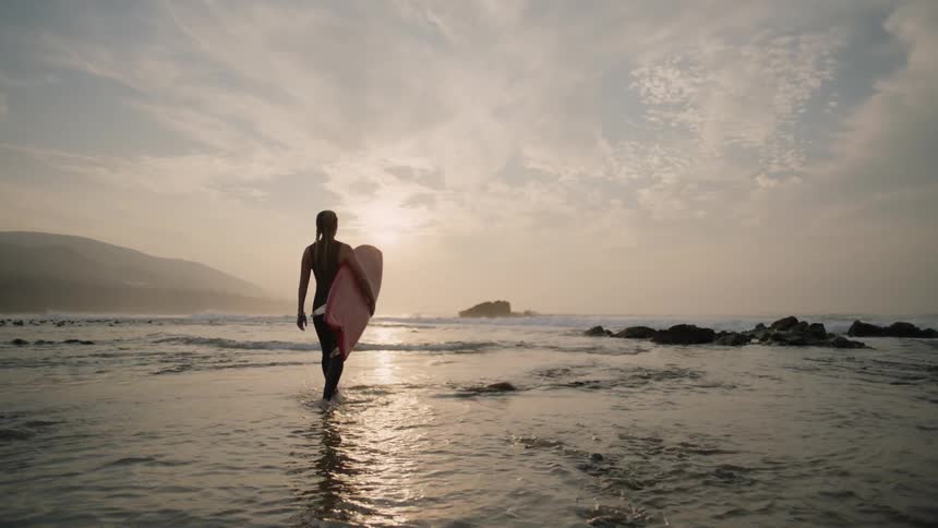 A woman holding a surf board walking out into the ocean as the sun reflects off the water as it starts to set