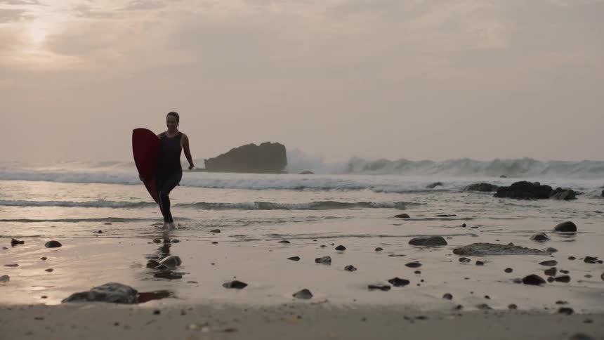 A woman running on the beach holding a surf board coming out of the water