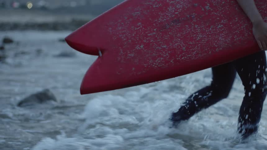 A woman barefoot wearing a wet suit walking across rocks and into the ocean carrying a red surf board
