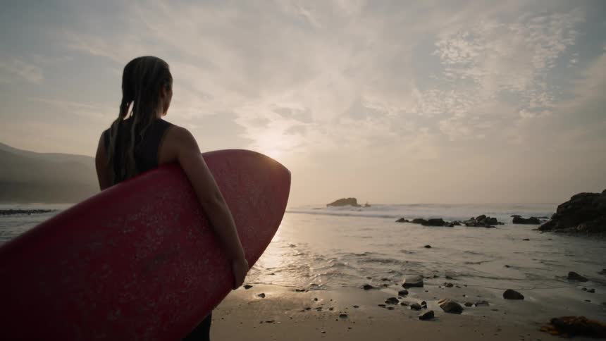 Young female surfer carries a red surfboard to the ocean at evening