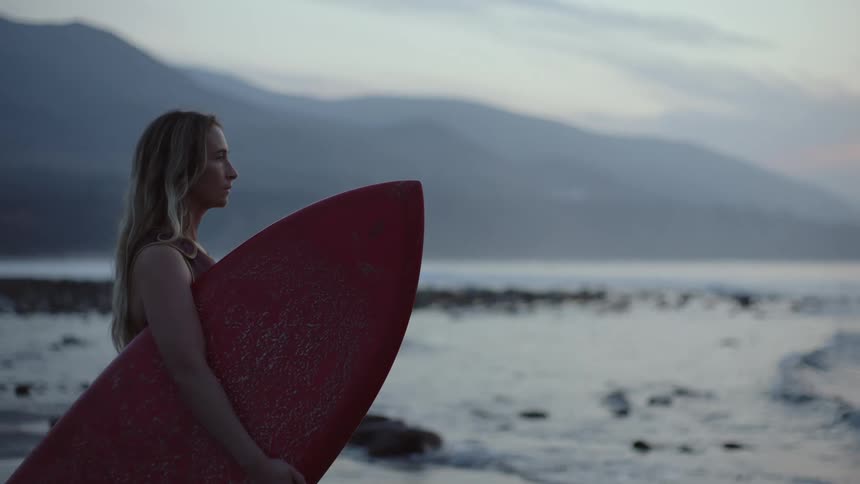 A woman stands by the water with her surfboard