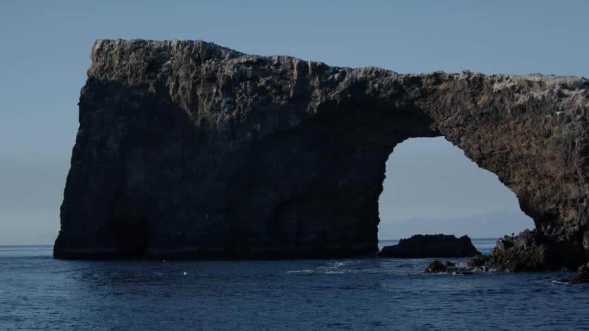 A drone view of a rock formation in the ocean with birds flying toward it