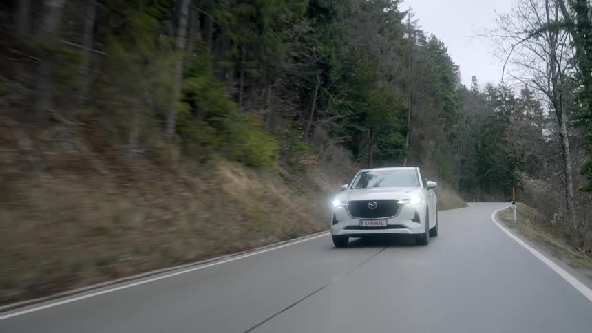 White vehicle drives along highway on mountain side covered by forest