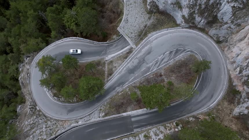 A white car drives along a winding mountain road before entering a tunnel