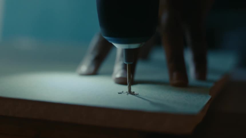 Person drills a hole on a plank of wood with a power tool on a dimly lit room