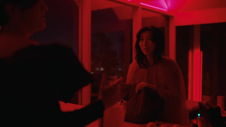 In room lit by red colored lighting one woman watches as another woman pours herself a drink