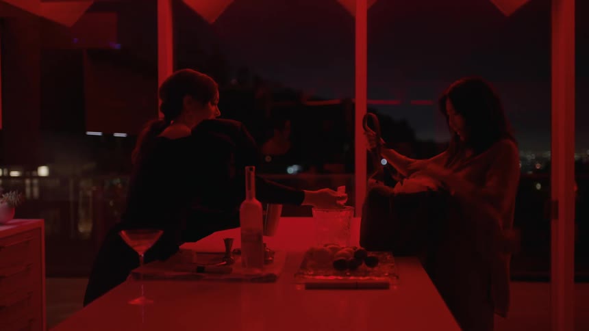 Two women walk into red lit room and one puts on wrap while the other starts making a drink