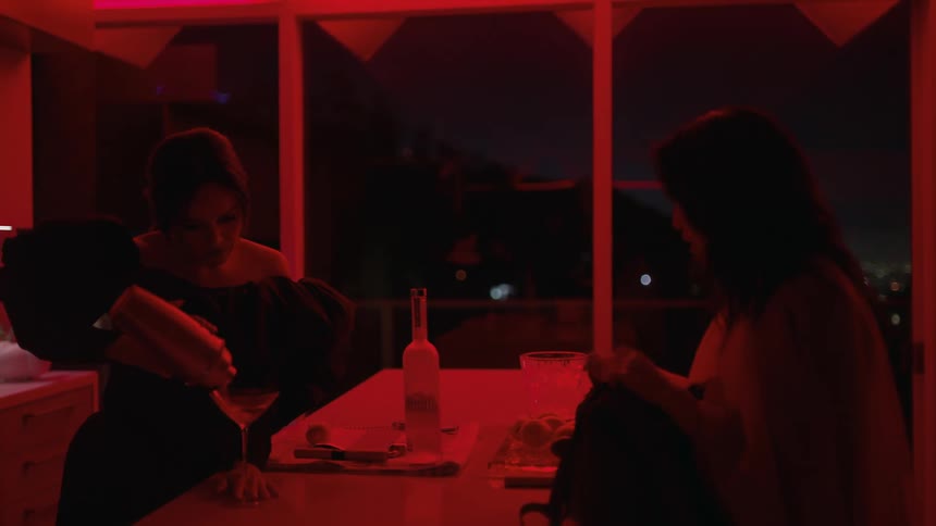 Two women standing in a red lit kitchen one preparing a drink while they talk