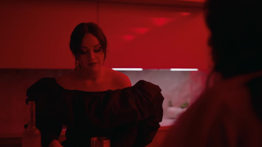 A woman in a black dress mixing an alcoholic drink in the kitchen as another woman walks around toward her