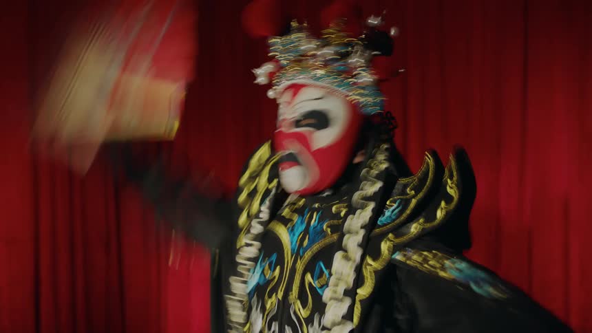 A person in costume and wearing a mask waving a fan around and dancing in front of a red curtain