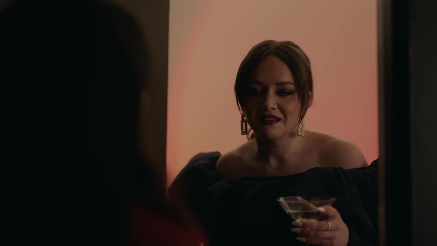 A woman in a black party dress holding a martini opens a door to talk to a friend in a dark room and invite her out