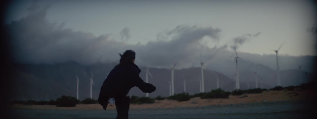 A woman is having difficulty walking against strong wind near a wind farm as dark storm clouds move overhead