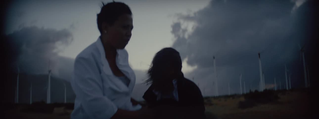 Mother tries to help daughter with her hair as they stand in road near wind farm with storm clouds in the sky