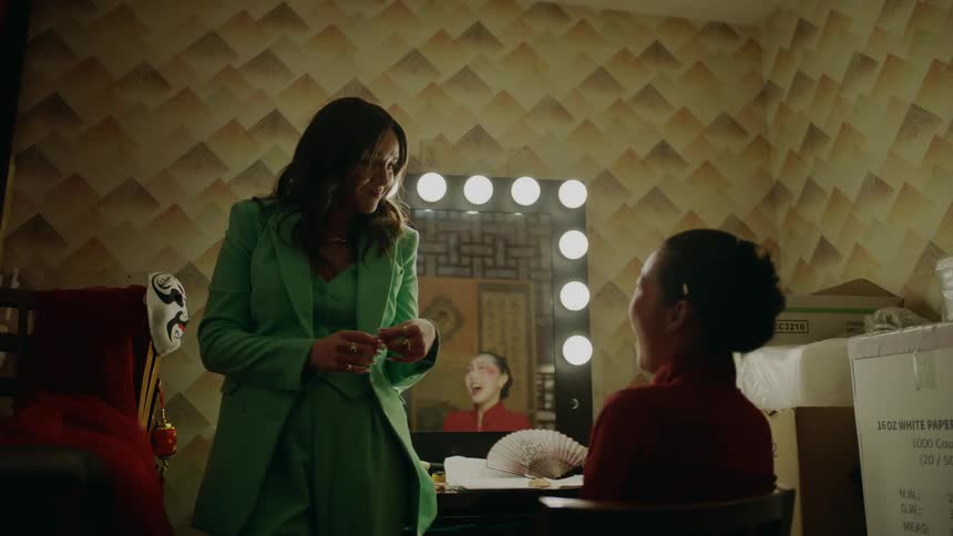 A woman in a green suit standing up talking to a woman in red sitting down as the woman standing puts something on the desk in front of the other woman