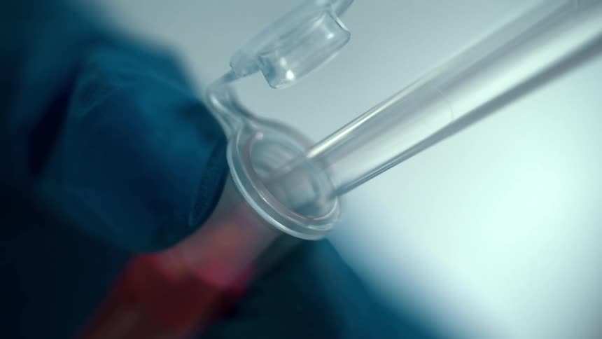 A close up of a persons hands wearing gloves holding a vile with liquid in it as a syringe draws the liquid out