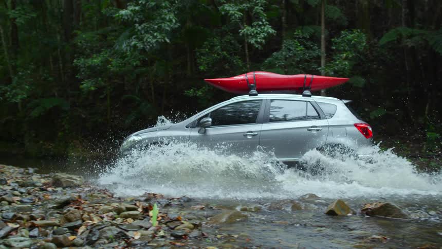 A small suv driving through a shallow stream with a red kayak on the top