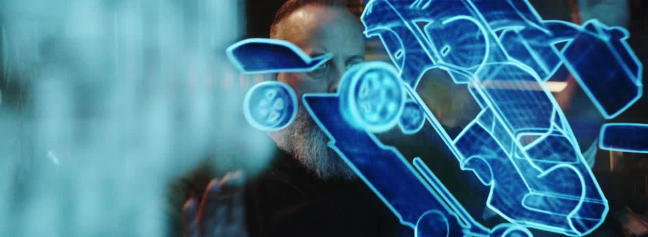 A man with a beard is moving a glowing holographic image of a car and it comes apart as he touches it