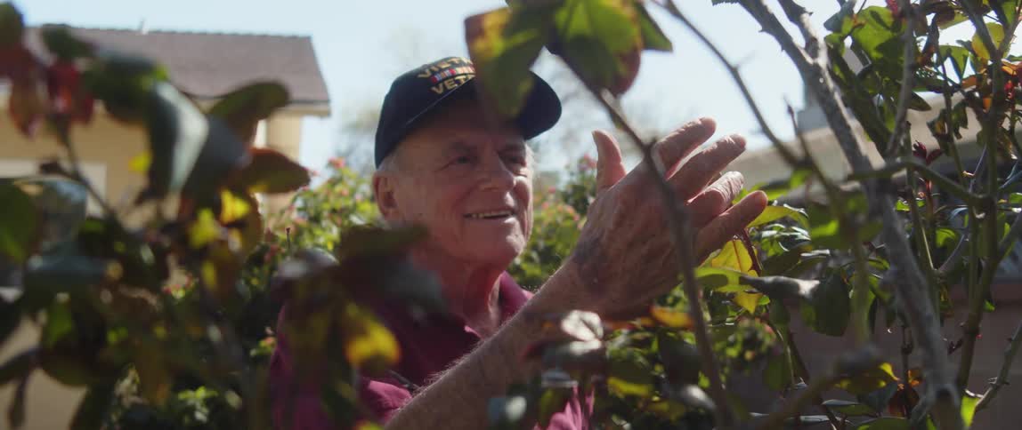 An elderly man in a cap smiles as he inspects a branch in the garden