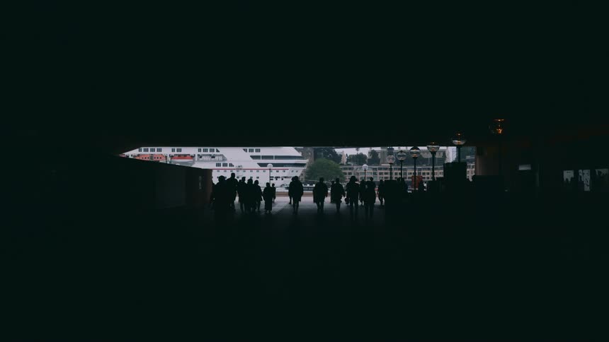 A crowd of people in a shadowed area walk toward a moored cruise ship