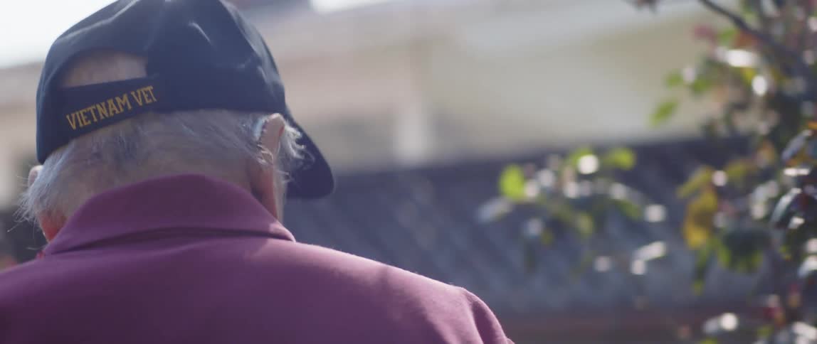 Old man with vietnam vet cap walking around outside his house inspecting plants and trees in his garden