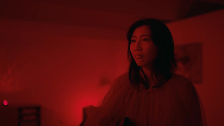 A woman stands completely still and looking scared in an empty room with a red light shining everywhere
