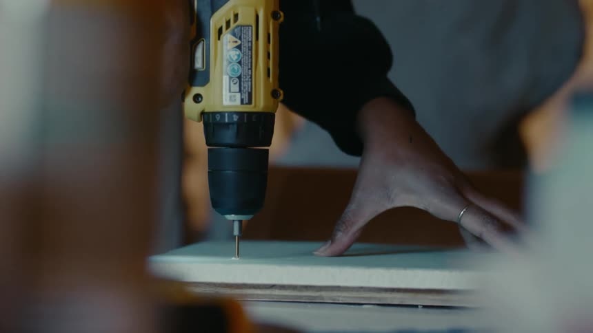 Carpenter uses a power drill to make a hole on a plank of wood in the dimly lit workshop