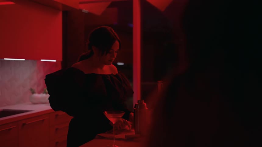 Woman prepares a cocktail while she and friend talk in kitchen lit by red light at night