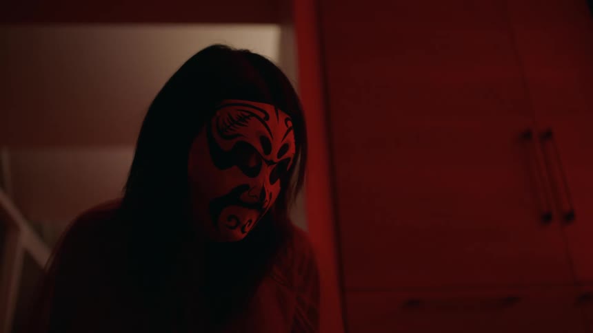 Close up of a woman wearing a chinese mask crouching on the floor of a room under red lights