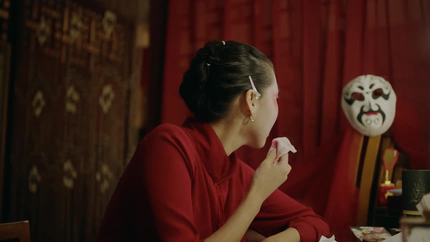 An asian woman sits in a dressing room at a table while removing her lipstick when a mask next to her moves on its own and is jarring to her