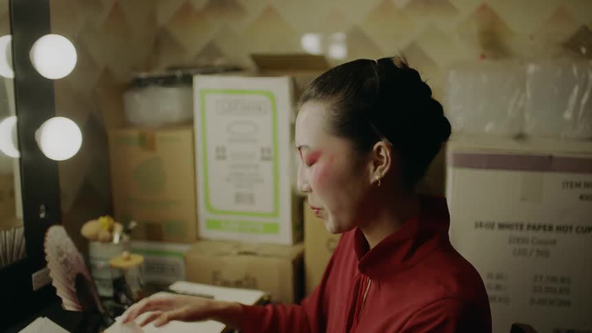 A woman sitting at a vanity in front of light holding a rag and looking around concerned