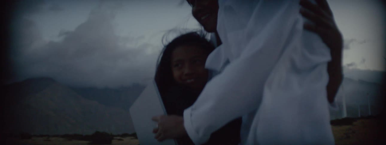 Two women with blouses waving in the wind hug a girl near wind turbines at evening