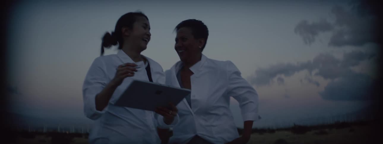 Mother with adult daughter walking together outside at sunset on a very windy day while holding a tablet and taking notes