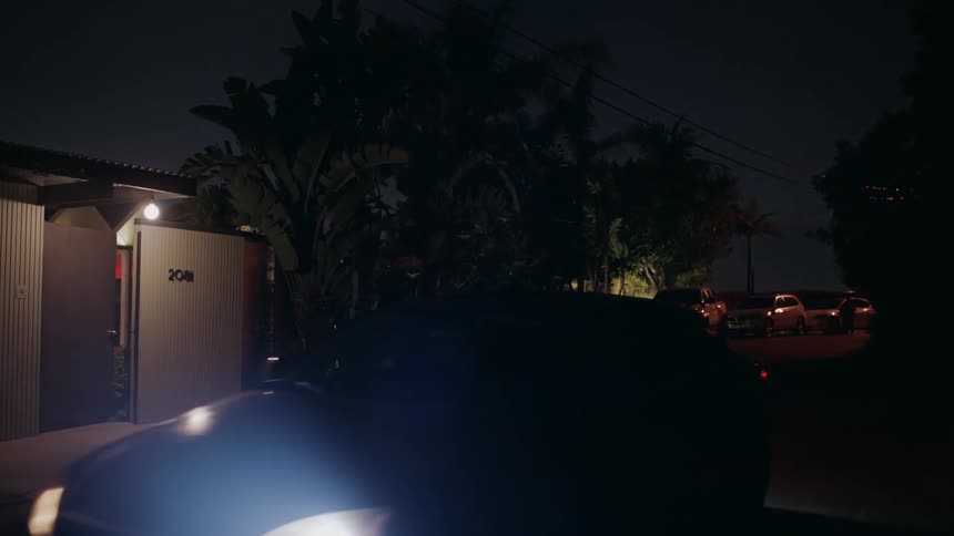 A car with headlights on parks in front of a house at night