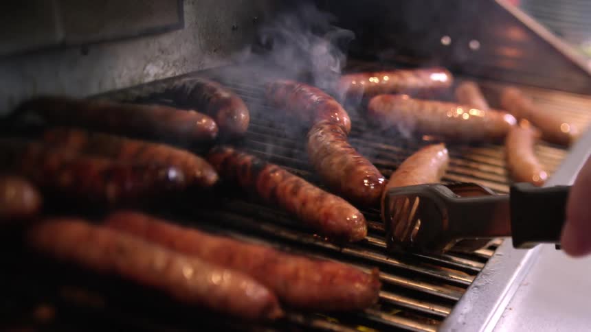 Close up of a person using tongs to turn sausages cooking over a grill