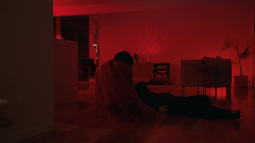 A person lying on the floor next to a person hunched over both in a dark room lit only by red accent light