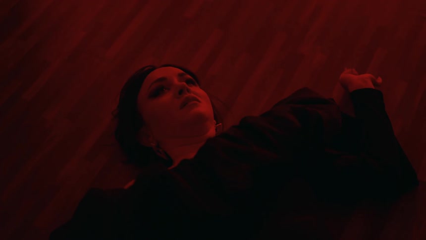 Woman wearing fashionable dress falls onto wooden floor in room lit by colored lighting and frowns tearfully