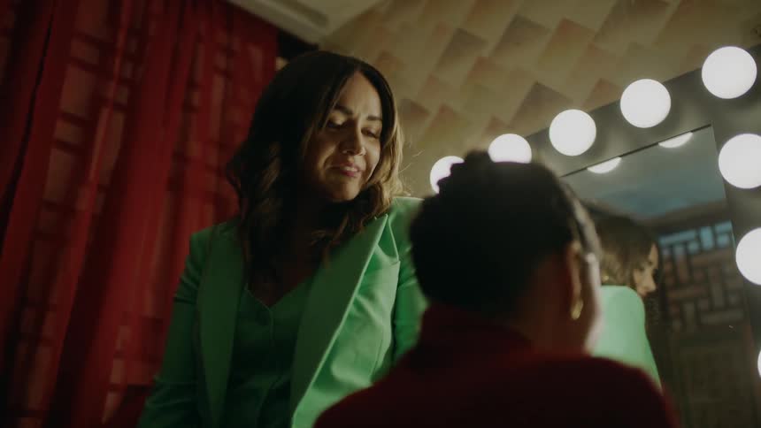 Two women talking in a dressing room one sitting down while the other walks over and stands in front of the woman sitting as they talk