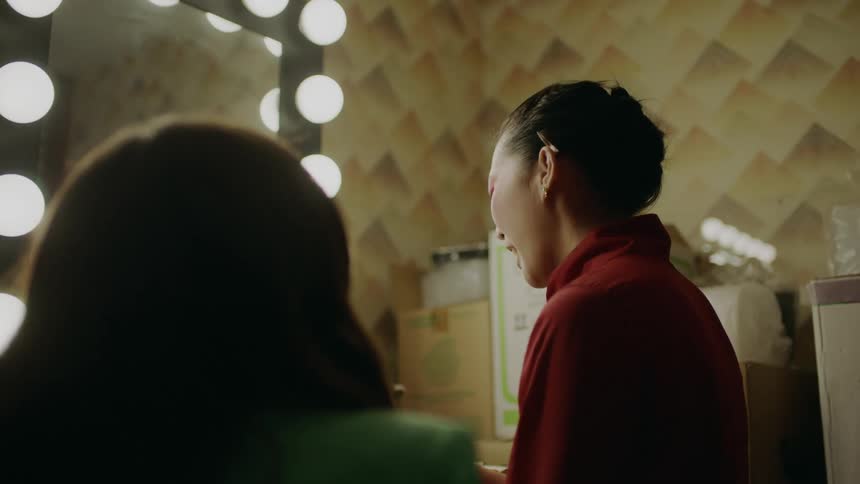 Woman sitting in front of mirror while applying makeup smiles while talking to another woman