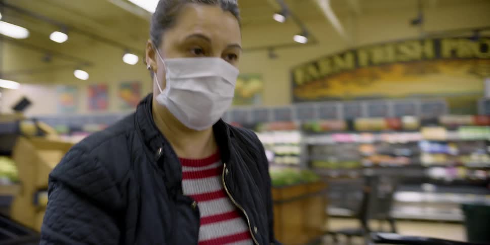 A woman wearing a mask and gloves places groceries into her shopping cart in a grocery store