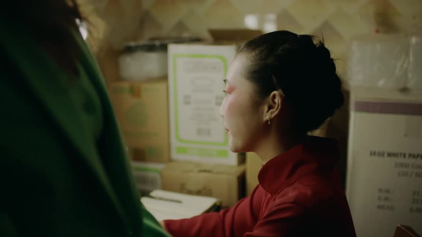 A woman sitting at a vanity smiling and talking to a woman walking up to her then looking down while the woman in green speaks to her